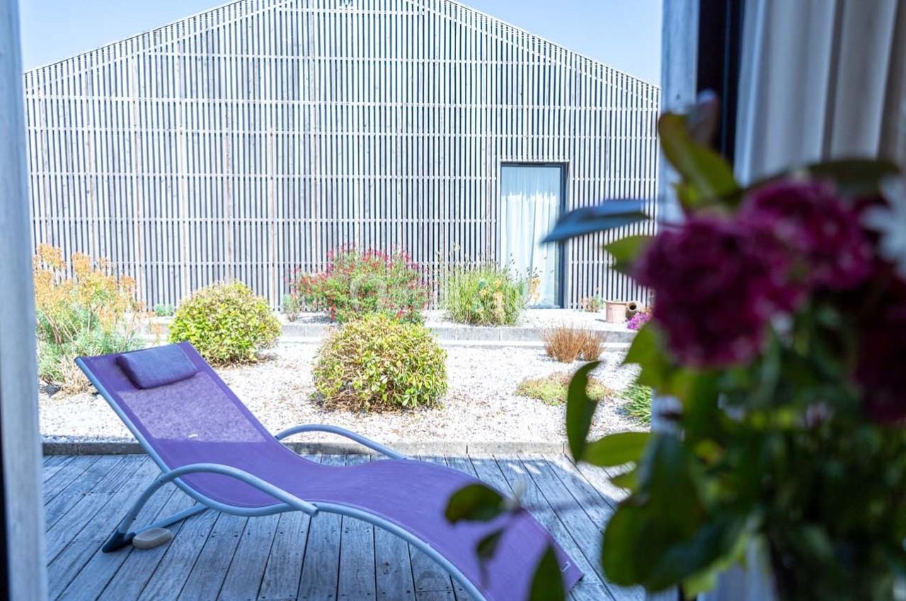 Chaise longue violette sur une terrasse en bois, avec vue sur un jardin et un bâtiment métallique en arrière-plan.