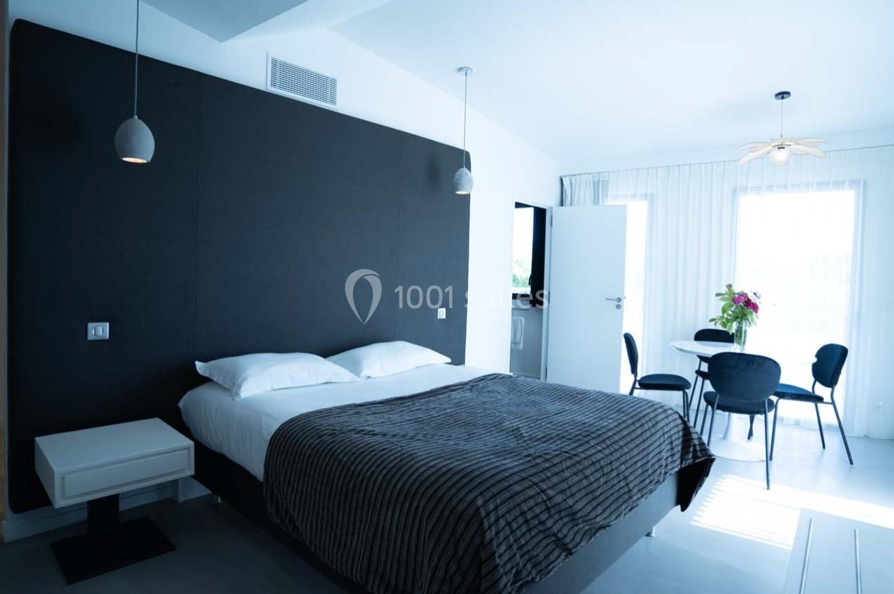 Chambre moderne avec un lit double, mur sombre, table avec chaises et lumière naturelle provenant d'une grande fenêtre.