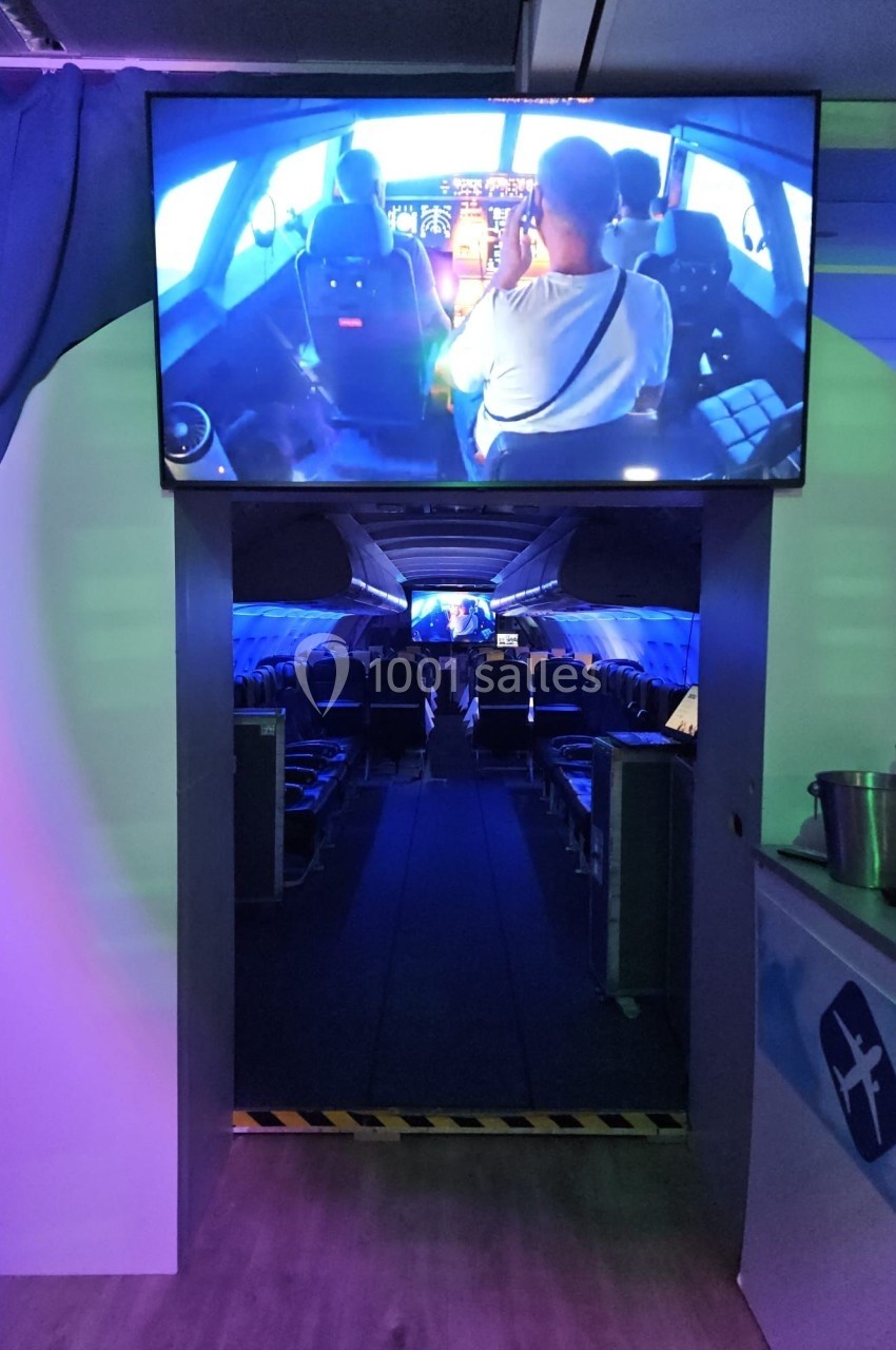 Vue intérieure d'un simulateur d'avion avec des sièges alignés et un écran montrant un cockpit en fonctionnement.