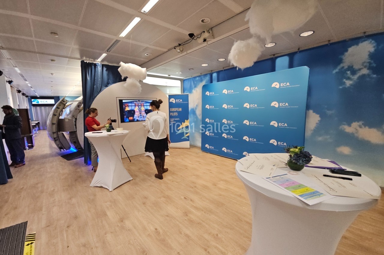 Salle d'exposition avec des stands, des écrans interactifs et des tables hautes, décorée de nuages suspendus.