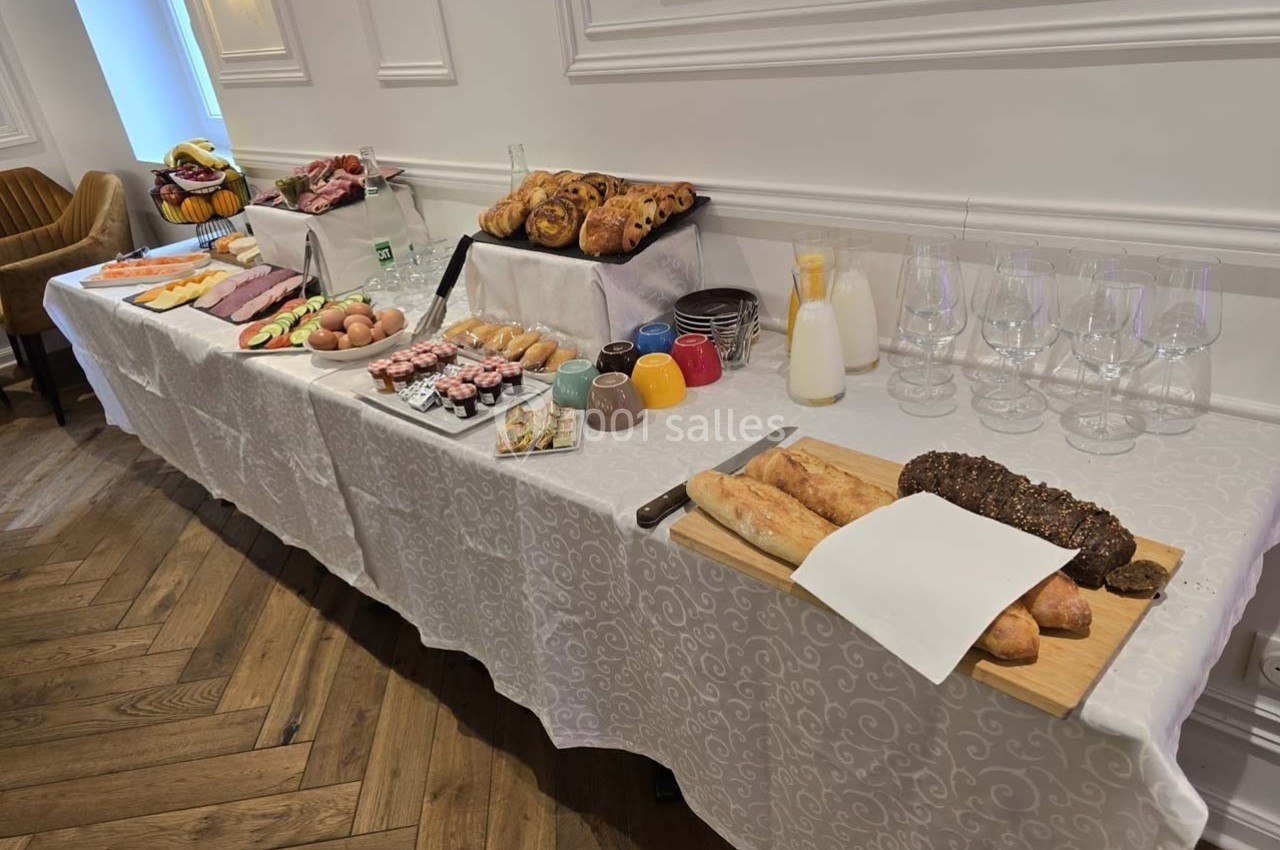 Buffet de petit-déjeuner avec viennoiseries, pains, fruits, charcuterie, œufs, jus et verres sur une table blanche.