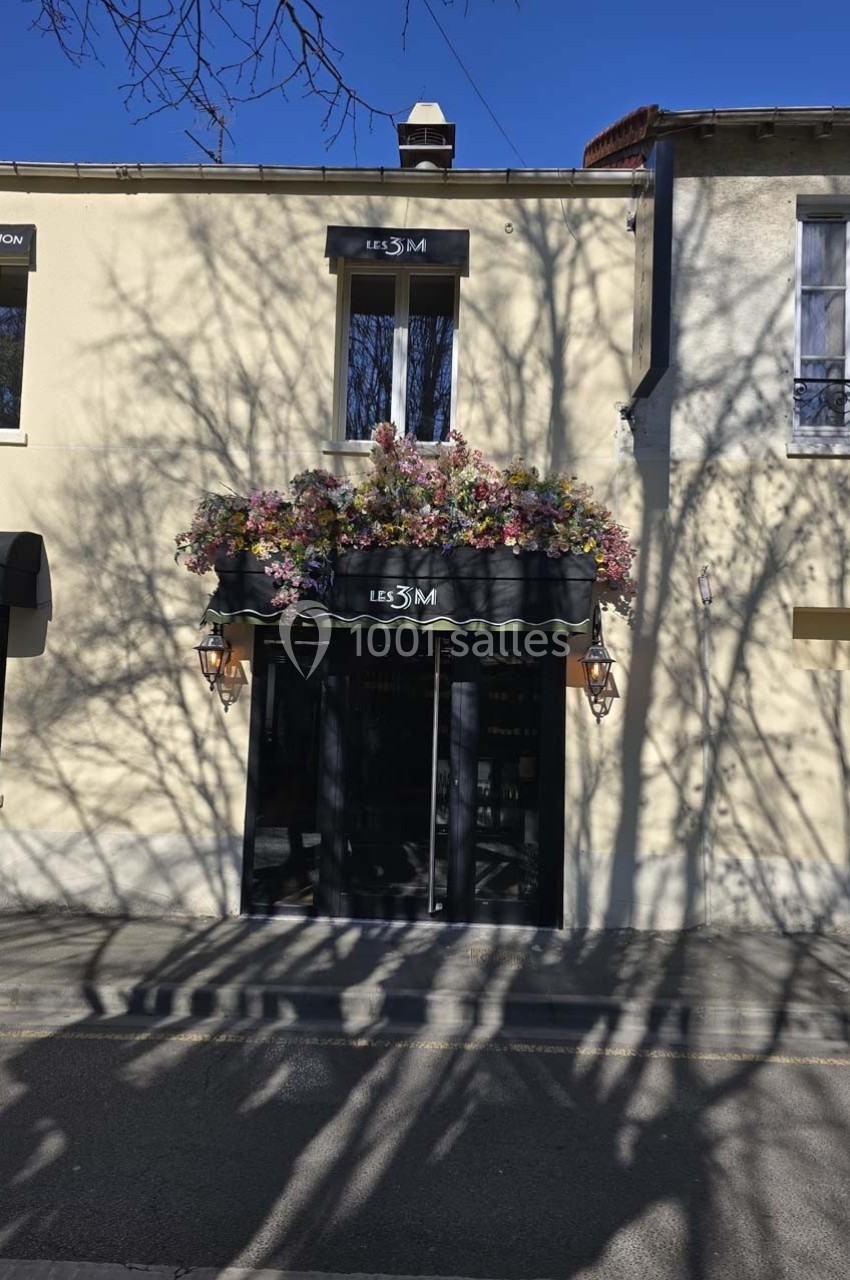 Façade d'un bâtiment avec une porte noire, une enseigne ’Les 3M’ et des fleurs colorées ornant le haut.