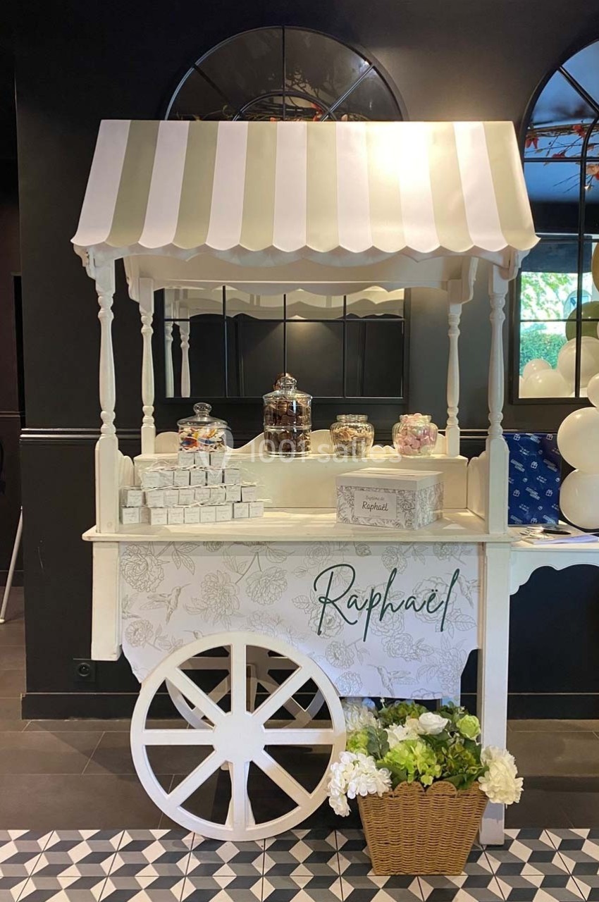 Chariot décoratif blanc avec friandises, bonbonnières et fleurs, placé dans un espace intérieur élégant.