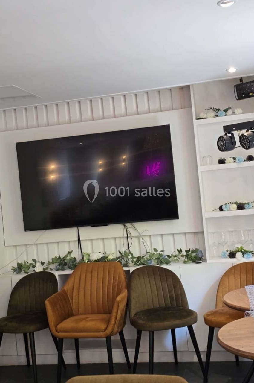 Télévision murale éteinte entourée d'étagères décorées de plantes, avec des chaises rembourrées alignées en dessous.