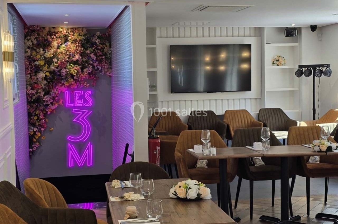 Salle de réception avec tables dressées, chaises modernes, mur floral et enseigne lumineuse ’Les 3M’.