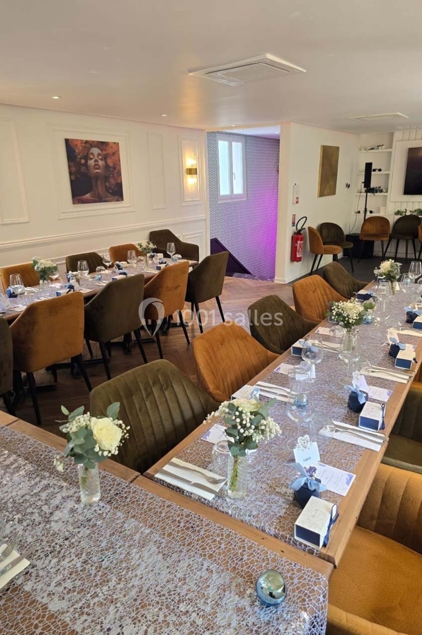 Salle élégamment décorée avec des tables dressées, chaises en velours, fleurs et lumière tamisée.