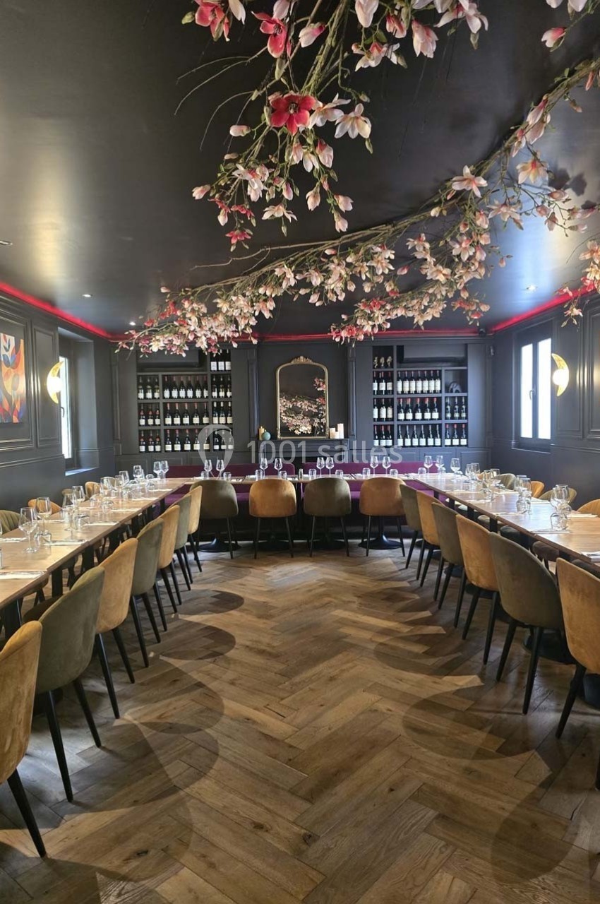 Salle de restaurant élégante avec tables dressées, parquet en bois, murs sombres et décoration florale suspendue.