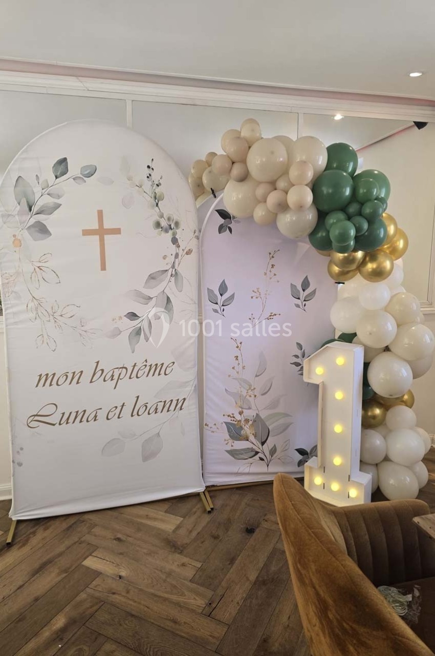 Décor de baptême avec arche de ballons blancs, verts et dorés, panneau floral et chiffre lumineux ’1’.