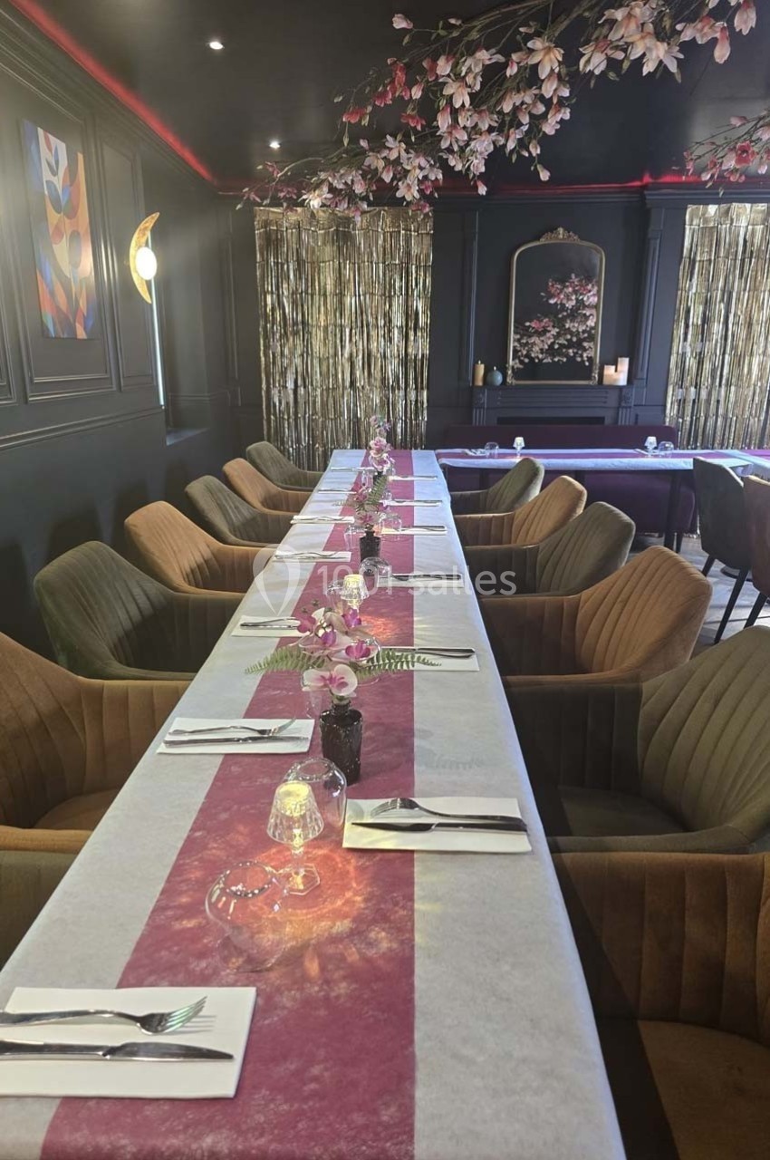 Salle de restaurant élégante avec une longue table dressée, décorée de fleurs et éclairée par une lumière tamisée.