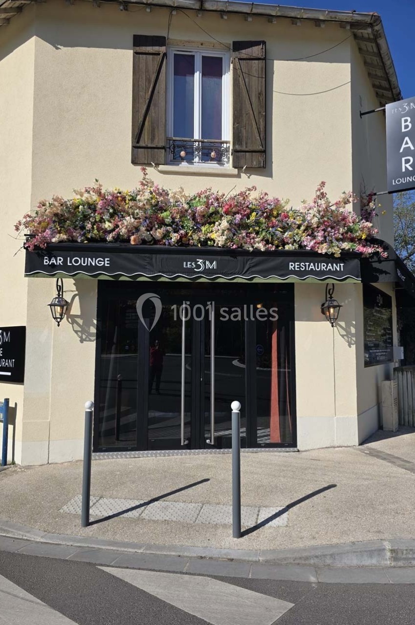 Façade d'un bar-restaurant avec une enseigne ’Les 3M’, terrasse fleurie et volets en bois sur une maison beige.