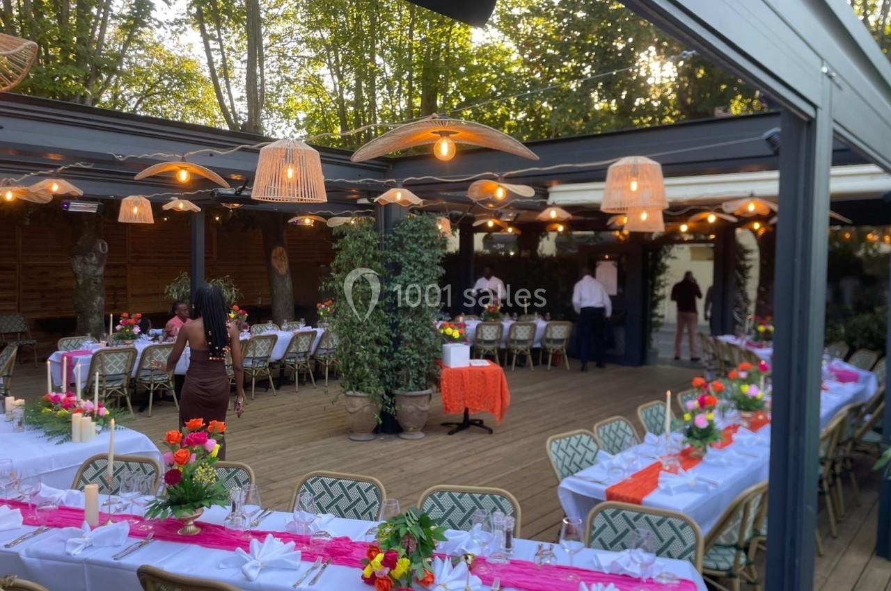 Terrasse décorée pour un événement, avec tables dressées, nappes blanches, fleurs colorées et éclairage suspendu.
