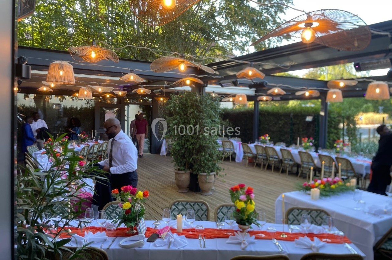 Terrasse décorée pour un événement, avec tables dressées, nappes blanches, fleurs colorées et guirlandes lumineuses.