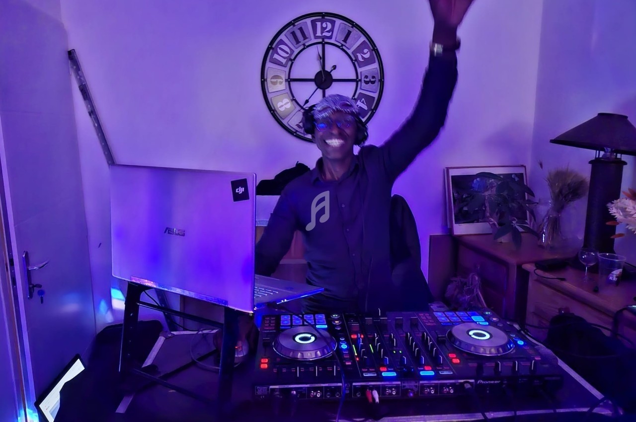 Un DJ mixant sur une table de mixage éclairée, levant une main dans une pièce à l'éclairage violet.
