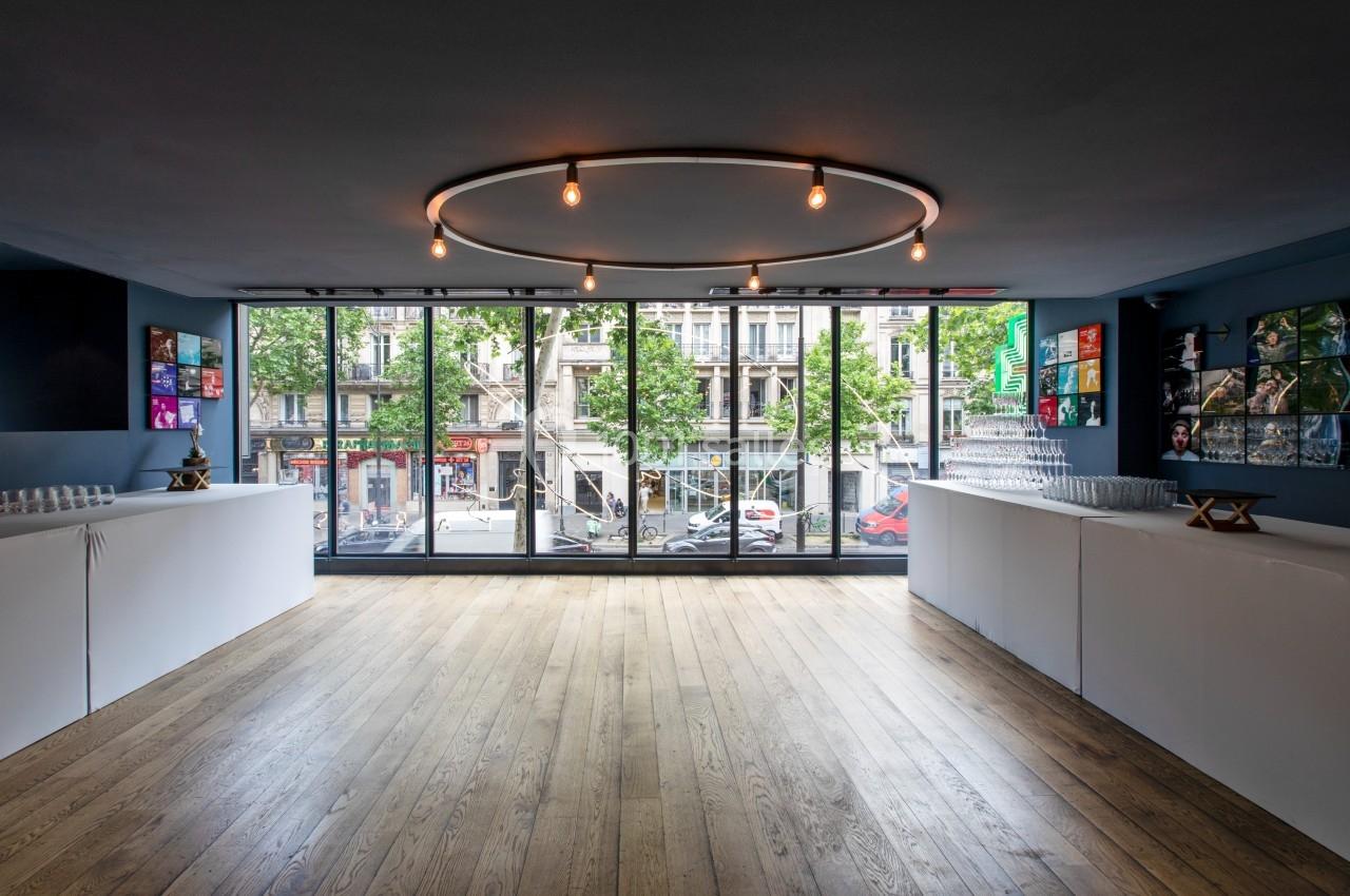 Salle lumineuse avec parquet, grandes baies vitrées donnant sur une rue urbaine, et comptoirs blancs alignés.