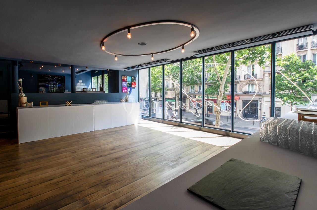 Salle lumineuse avec parquet en bois, grande baie vitrée donnant sur une rue urbaine et comptoir blanc moderne.