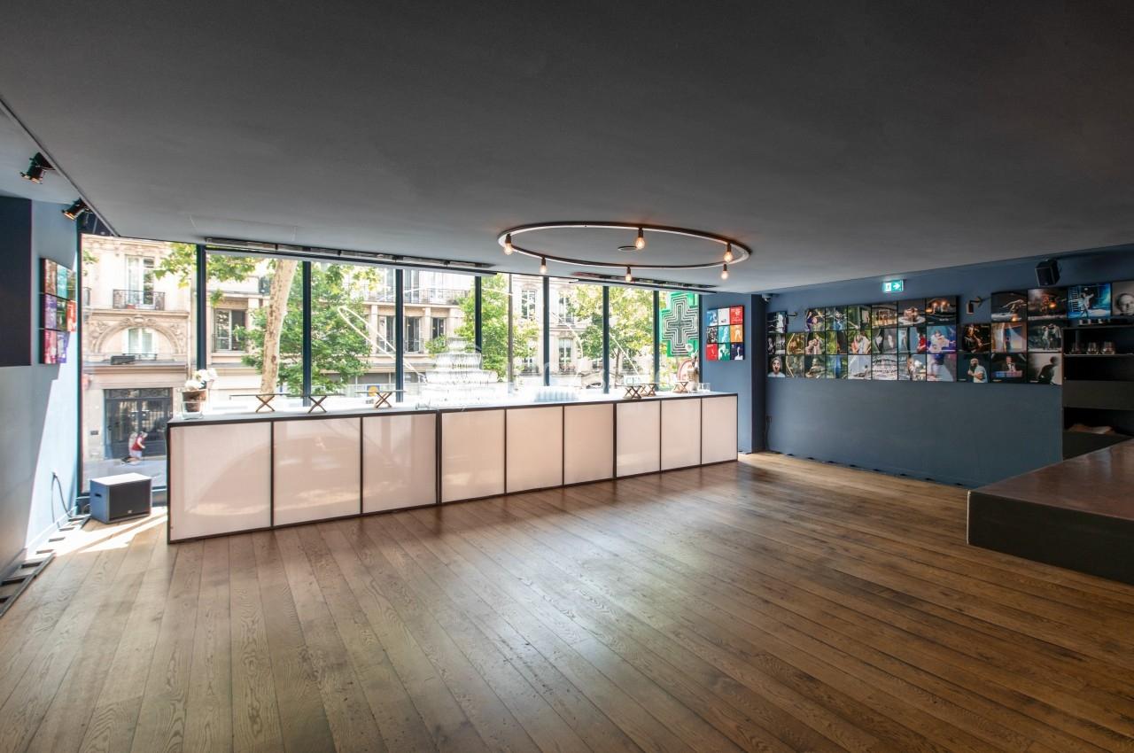 Salle lumineuse avec parquet en bois, grandes baies vitrées donnant sur la rue et étagères décorées sur le mur.