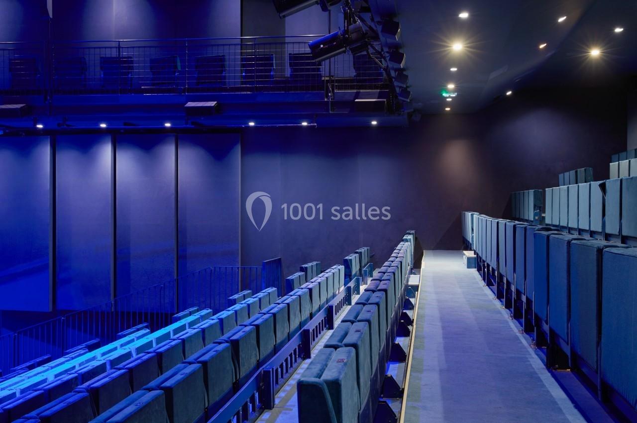 Salle de spectacle moderne avec rangées de sièges bleus, éclairage tamisé et passerelle en hauteur.