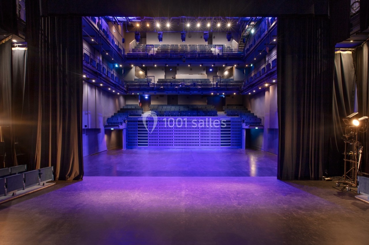 Vue depuis la scène d'une salle de théâtre vide, éclairée par des projecteurs avec des rangées de sièges en arrière-plan.