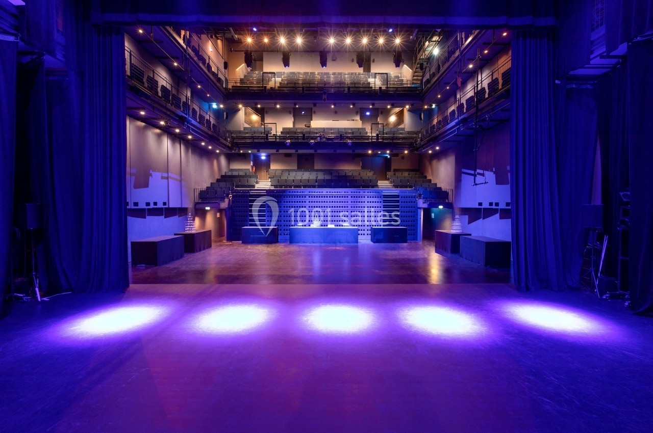 Vue depuis une scène éclairée sur une salle de spectacle vide avec gradins et balcons sous un éclairage violet.