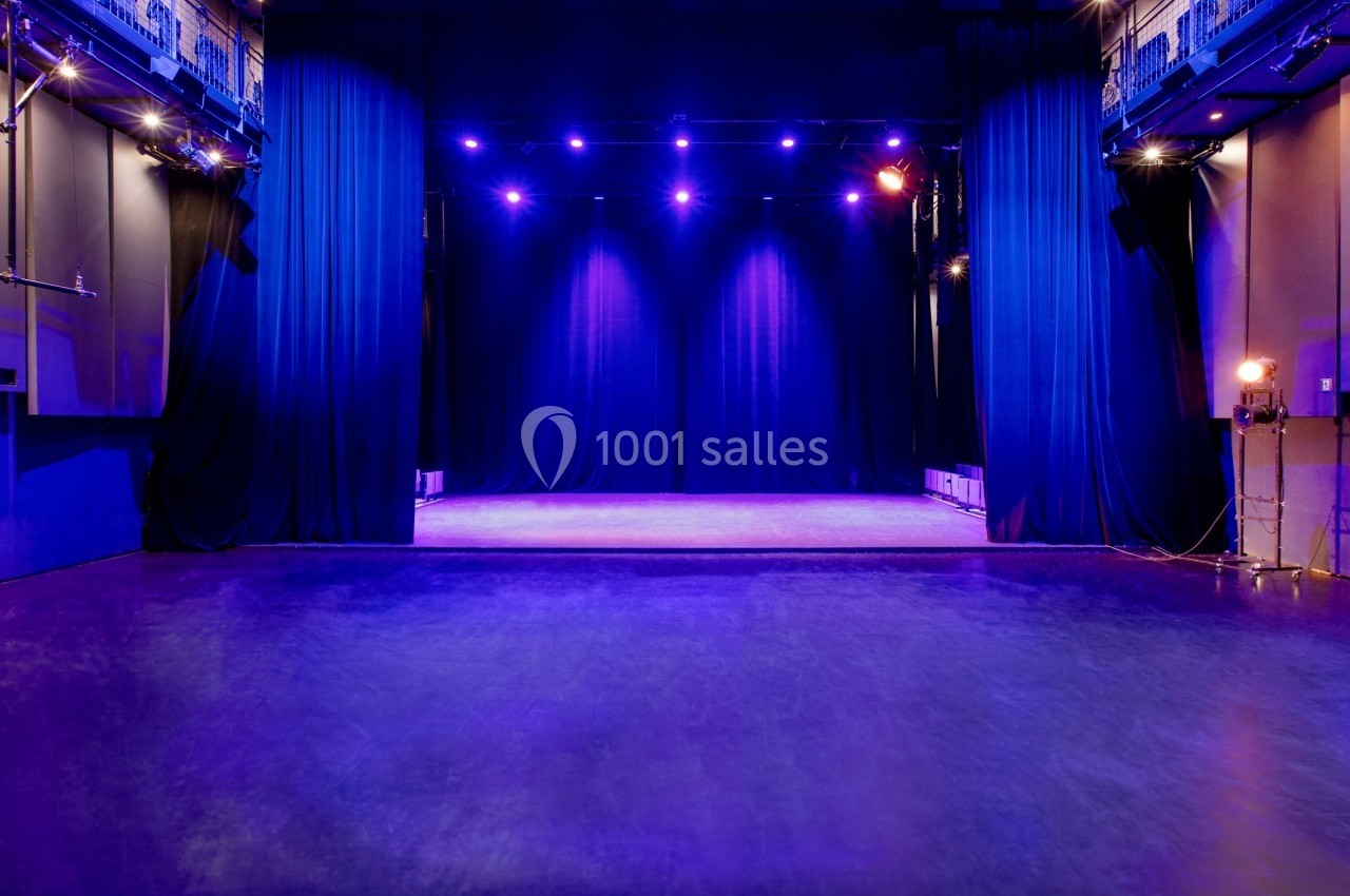 Scène de théâtre vide éclairée par des projecteurs violets, avec rideaux noirs et sol en parquet sombre.