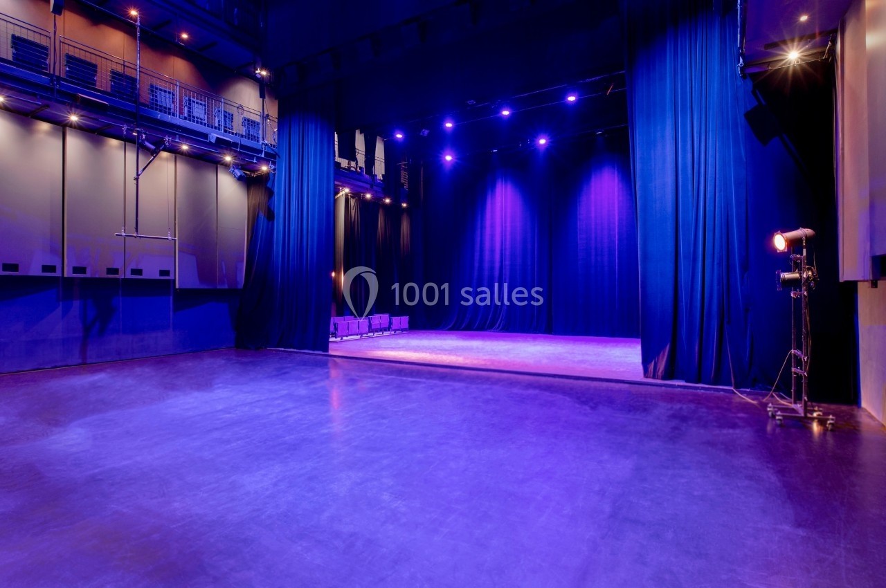 Salle de spectacle vide avec scène éclairée par des projecteurs violets, entourée de rideaux noirs.