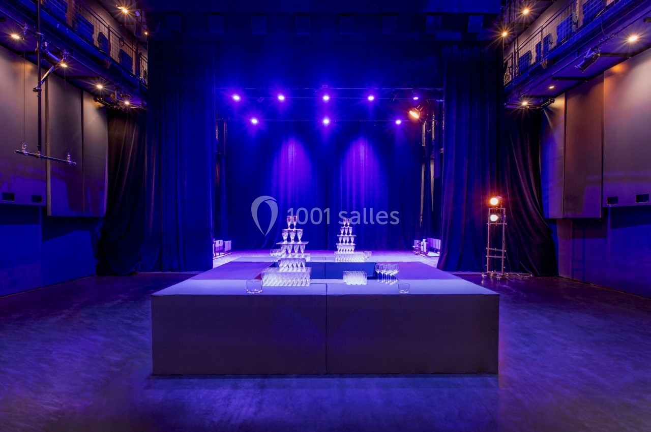 Salle de spectacle avec éclairage violet, scène en arrière-plan et tables décorées de pyramides de verres au premier plan.