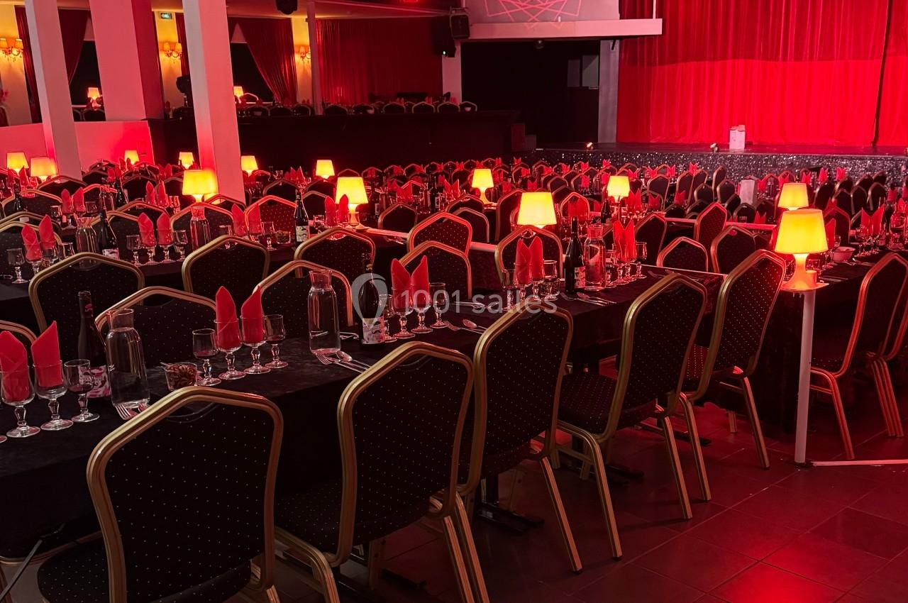 Salle de spectacle avec tables dressées, nappes noires, serviettes rouges et lampes allumées, ambiance tamisée.