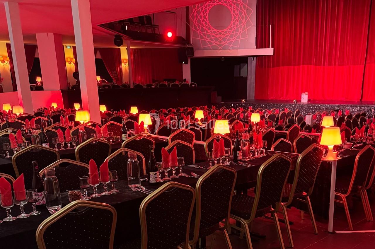 Salle de spectacle avec tables dressées, nappes noires, serviettes rouges et lampes diffusant une lumière tamisée.