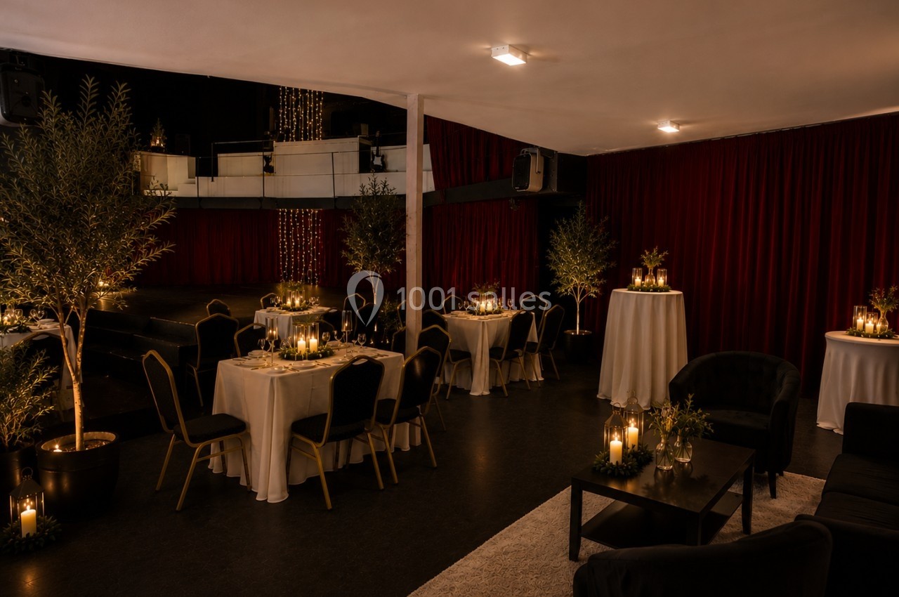 Salle décorée pour un événement, avec tables dressées, bougies, plantes et rideaux rouges en arrière-plan.