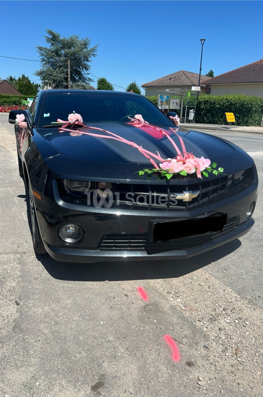 Voiture noire de type coupé Chevrolet décorée de rubans et de roses roses sur le capot et les rétroviseurs, garée sur une route ensoleillée. Let me check length: roughly 150 characters, too long. Need ≤125. "Coupé Chevrolet noir décoré de rubans et de