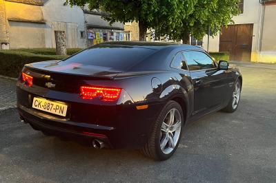 Vue de l'intérieur d'une voiture Chevrolet avec volant, tableau de bord et console centrale, devant un paysage urbain.