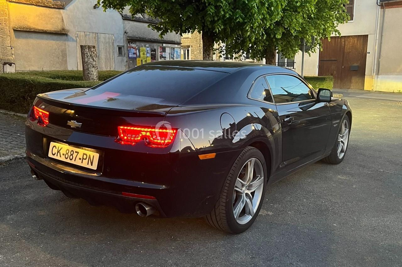 Voiture noire garée sur une place pavée, éclairée par une lumière douce au crépuscule, avec des bâtiments en arrière-plan.
