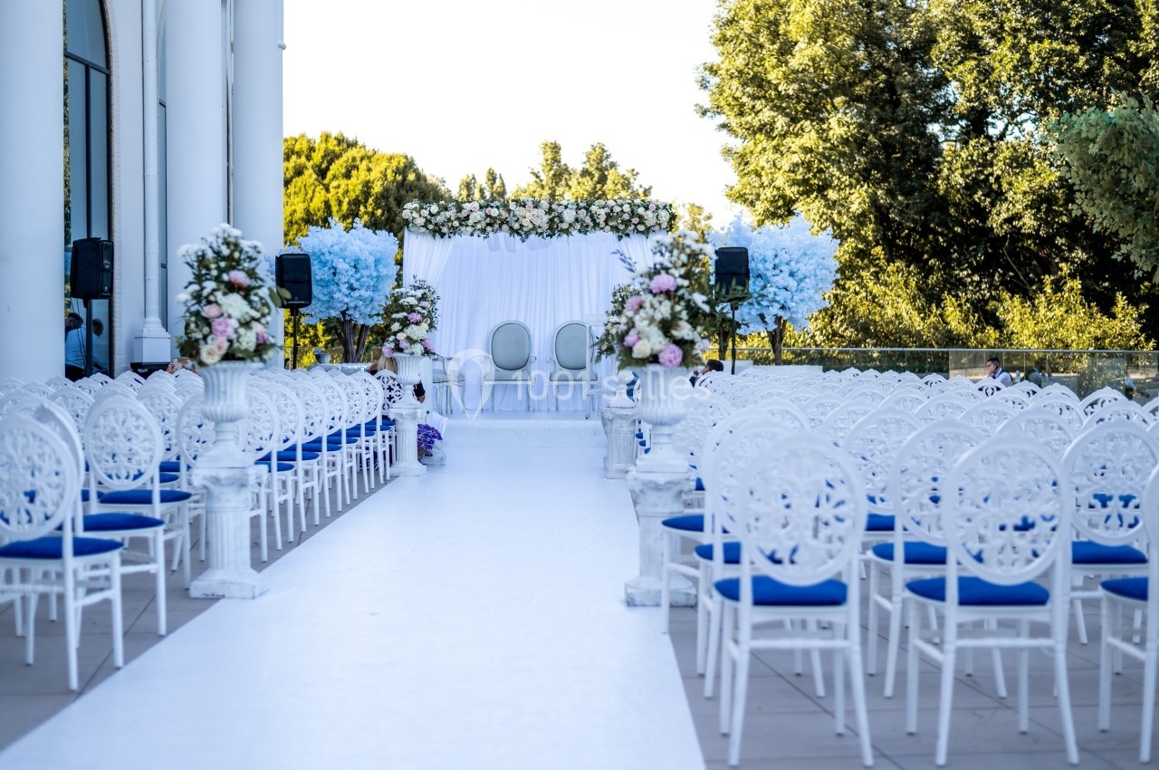 Allée centrale bordée de chaises blanches et décorations florales menant à une arche de mariage en extérieur.
