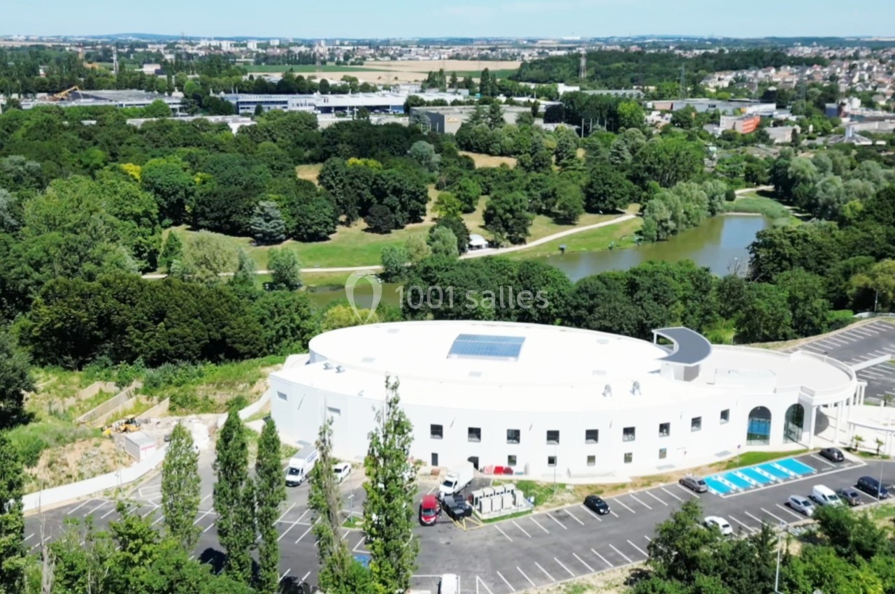 Vue aérienne d'un bâtiment blanc circulaire entouré d'arbres, avec un parking et un plan d'eau à proximité.