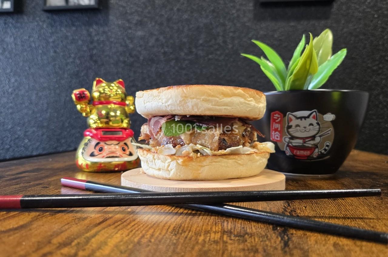Un burger garni posé sur une table en bois, accompagné de baguettes, d'une plante et de décorations japonaises.