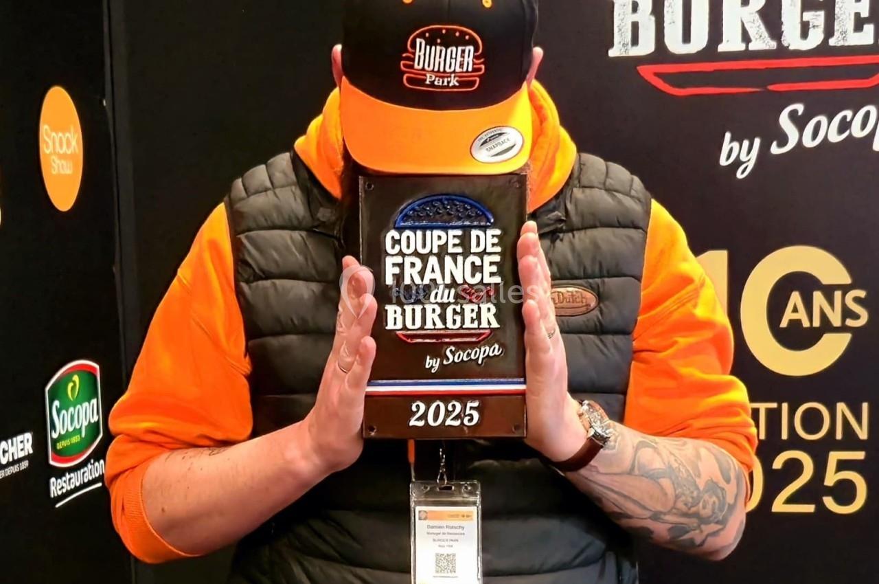 Un homme tenant un trophée marqué ’Coupe de France du Burger 2025’ devant son visage, portant une casquette et un sweat…