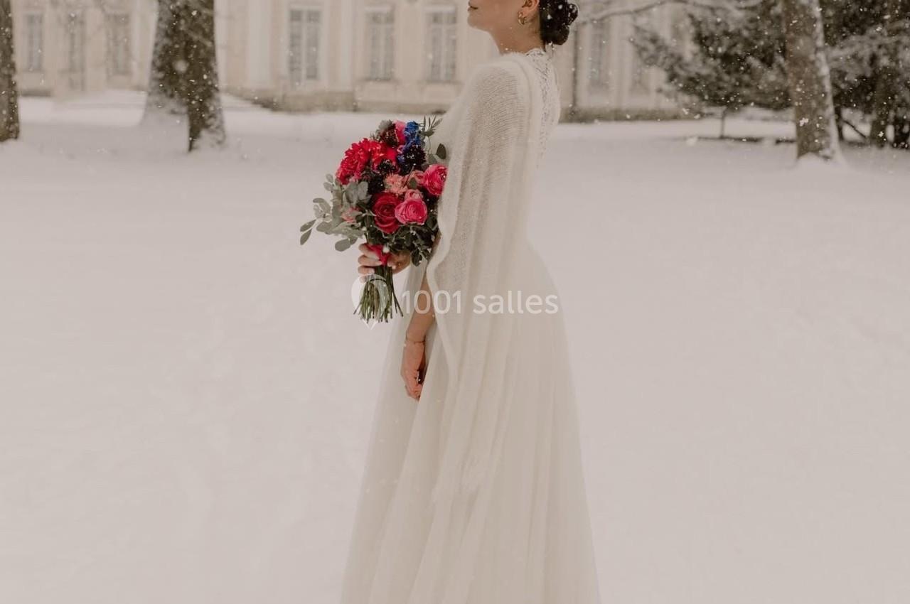 Une femme en robe de mariée blanche tient un bouquet coloré dans un parc enneigé devant un bâtiment ancien.