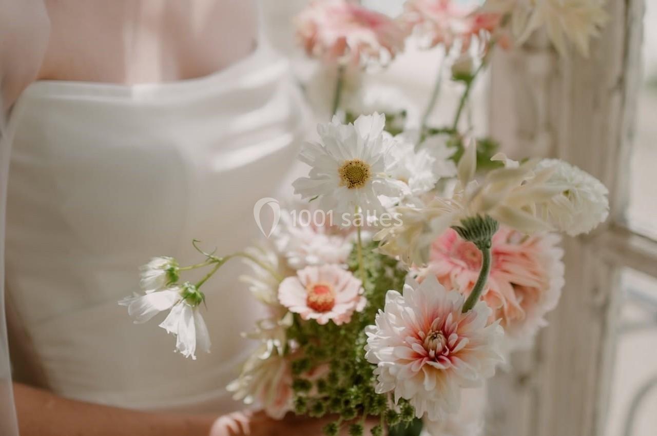 Femme en robe blanche tenant un bouquet de fleurs pastel devant une fenêtre lumineuse.