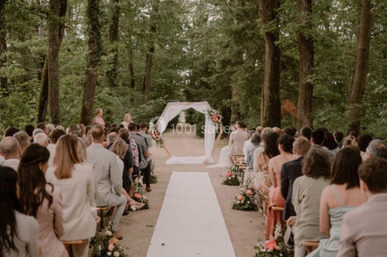 Cérémonie de mariage en plein air dans une forêt, avec une allée centrale bordée de fleurs et des invités assis des deux…