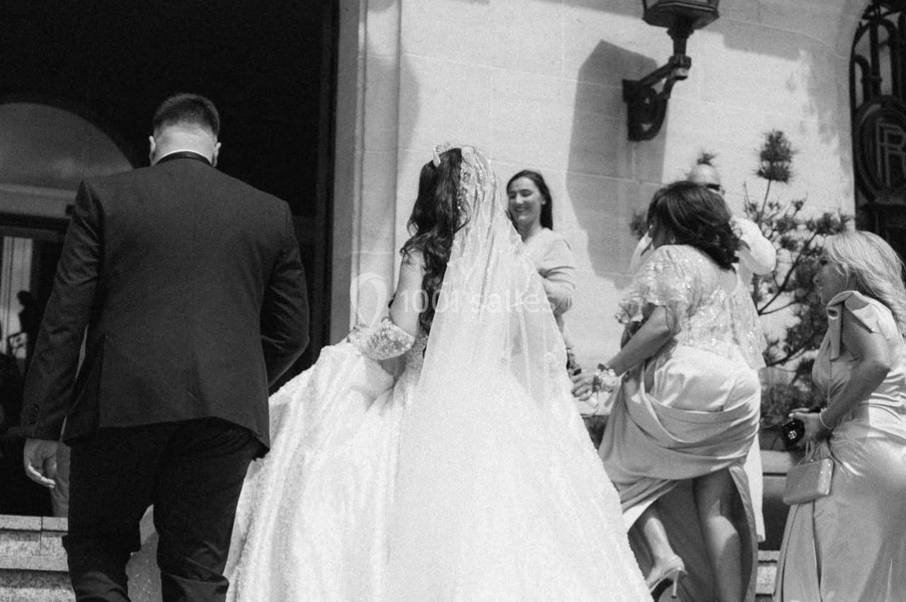 Un couple monte des marches, la mariée en robe blanche et voile, entourée d'invités élégamment vêtus.