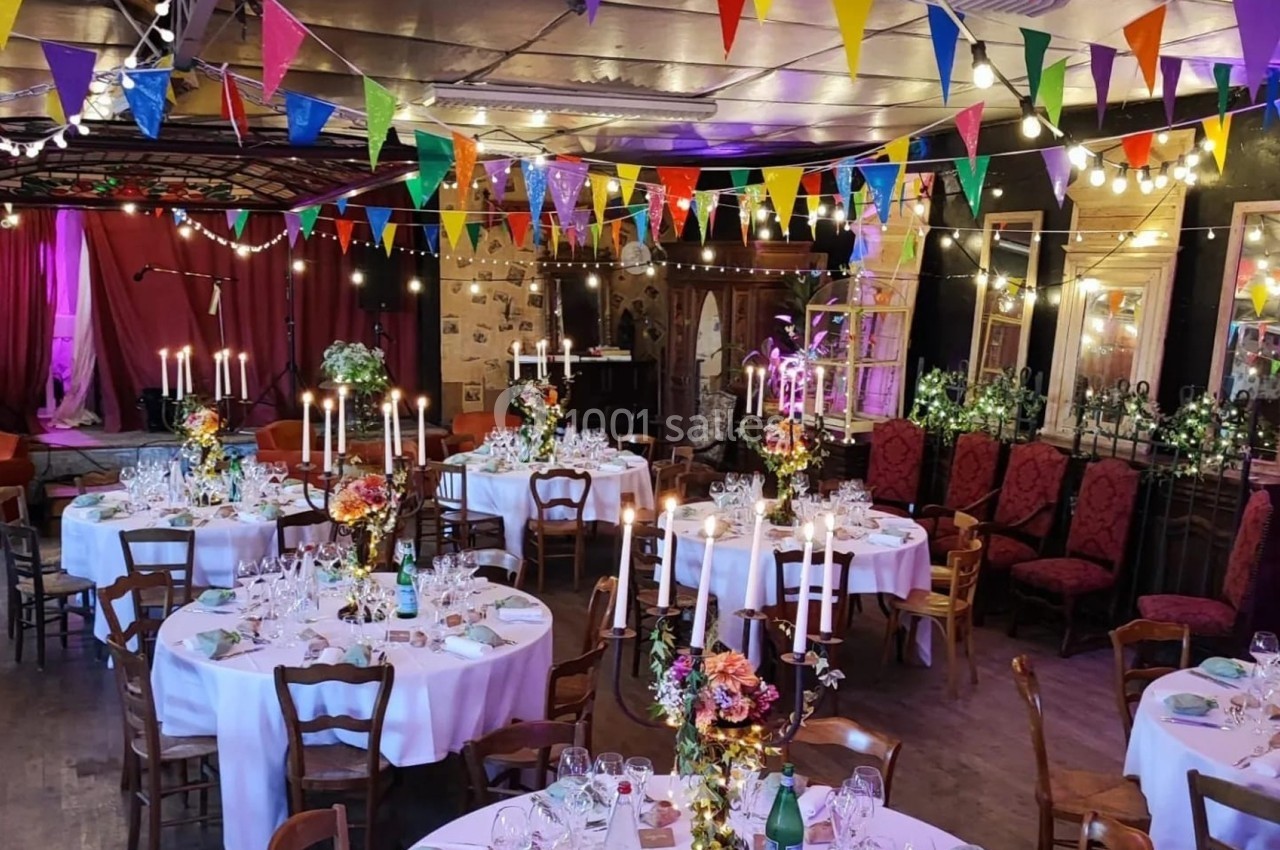 Salle décorée pour un événement, avec tables rondes dressées, chandeliers, guirlandes colorées et éclairage chaleureux.