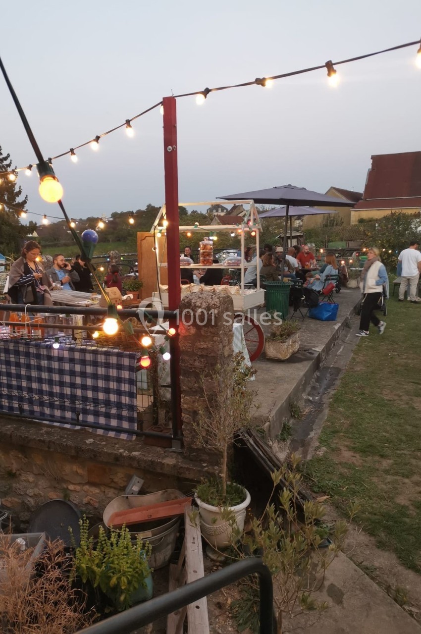 Personnes rassemblées en plein air autour de stands et tables décorées, sous des guirlandes lumineuses au crépuscule.