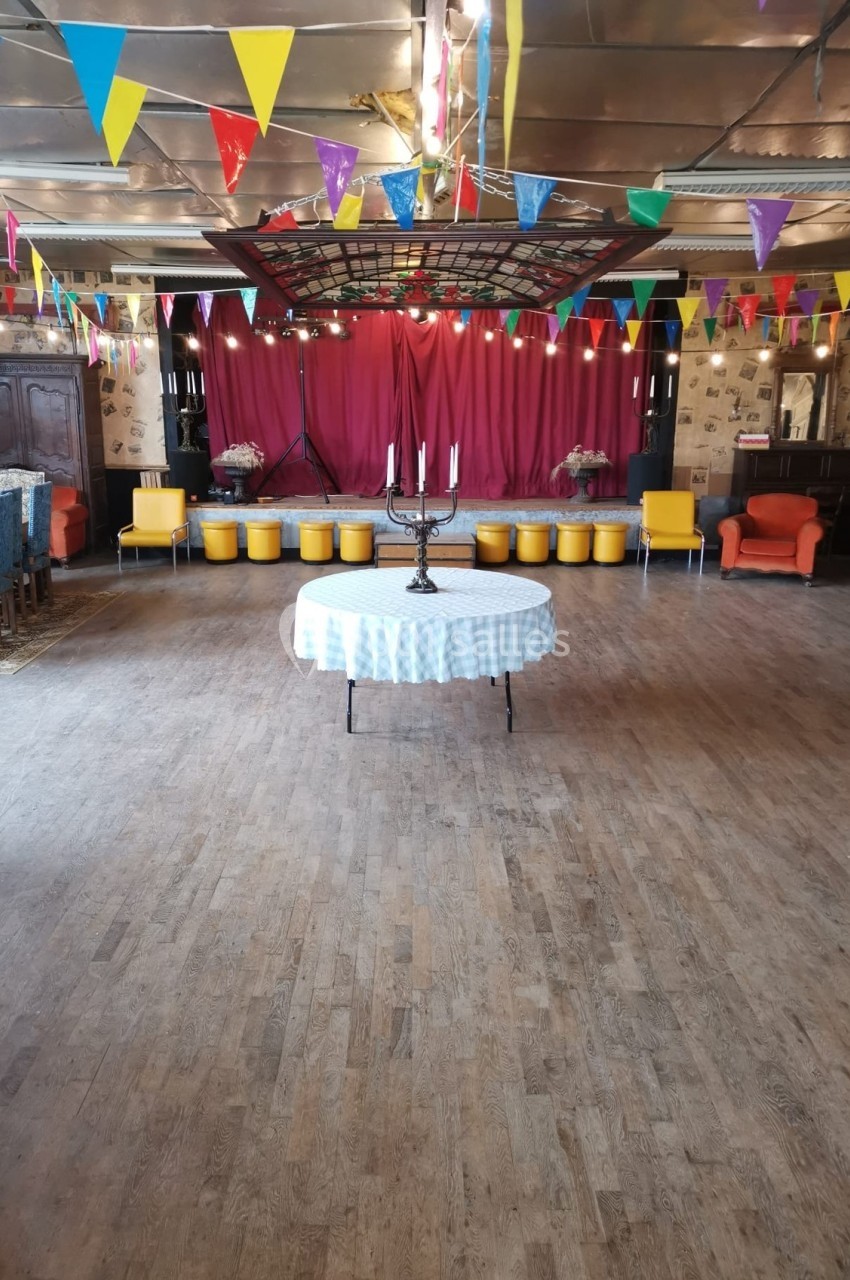 Salle décorée avec des guirlandes colorées, une table ronde au centre et une scène avec rideaux rouges en arrière-plan.