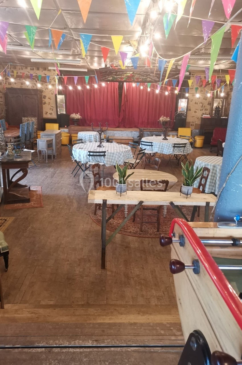 Salle décorée avec des guirlandes colorées, tables couvertes de nappes à carreaux, scène en fond avec rideaux rouges.