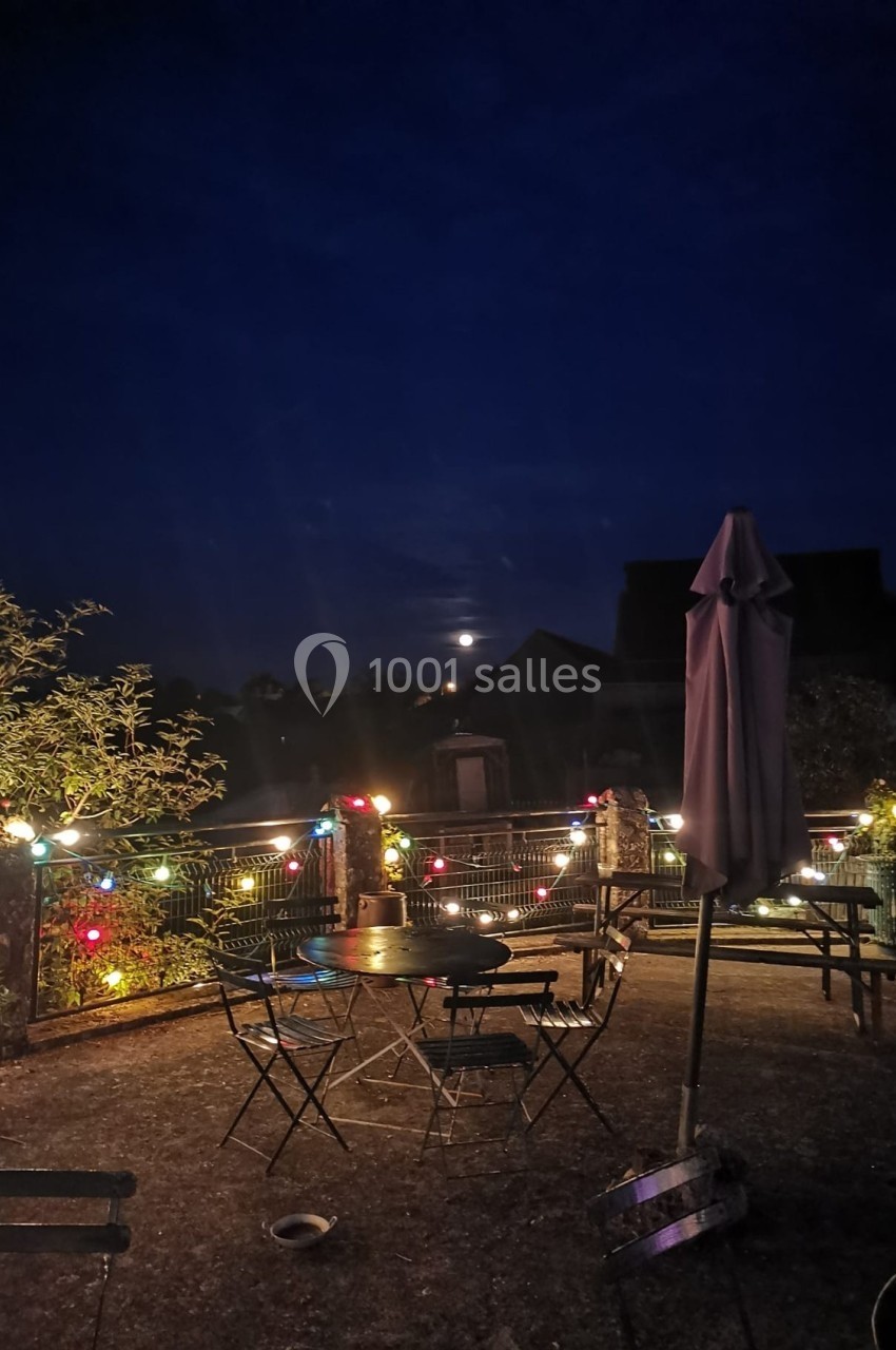 Terrasse extérieure éclairée par des guirlandes lumineuses colorées sous un ciel nocturne avec une pleine lune.