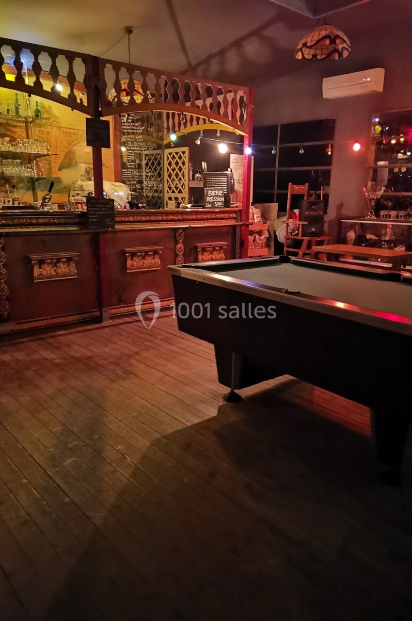 Salle de bar avec comptoir en bois, table de billard au premier plan et éclairage tamisé.