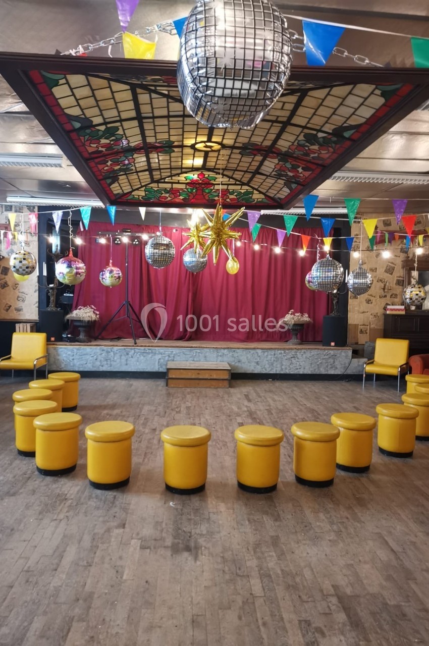 Salle décorée pour un événement avec guirlandes, boules à facettes, tabourets jaunes et une scène en fond.