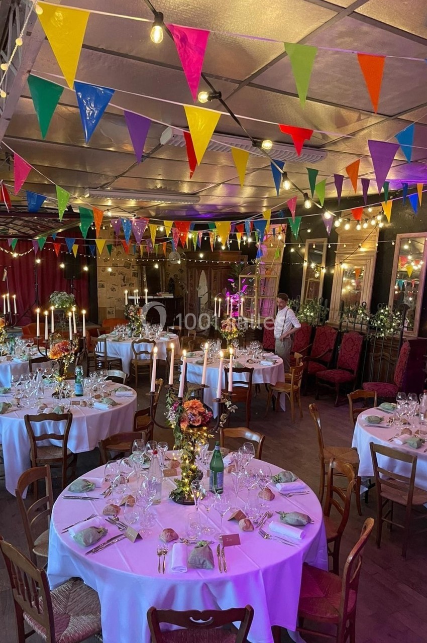 Salle décorée pour un événement avec tables dressées, chandeliers, guirlandes lumineuses et fanions colorés suspendus.