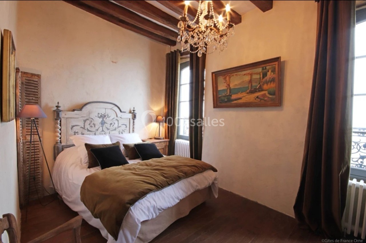 Chambre rustique avec lit double, poutres apparentes, lustre, tableau mural et fenêtre donnant sur l'extérieur.