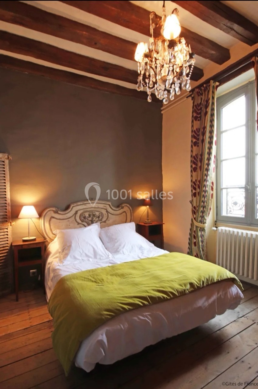 Chambre rustique avec lit double, poutres apparentes, lustre, rideaux colorés et lumière naturelle d'une fenêtre.